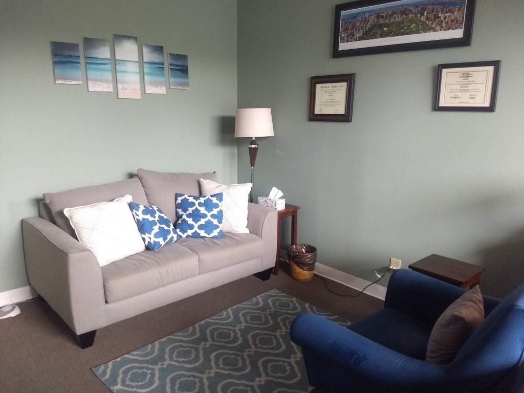 Image description: Ventura's office's carpeted therapy room featuring light green walls, a gray loveseat couch and a blue accent chair. The office is shown with natural lighting and feels warm and inviting.
