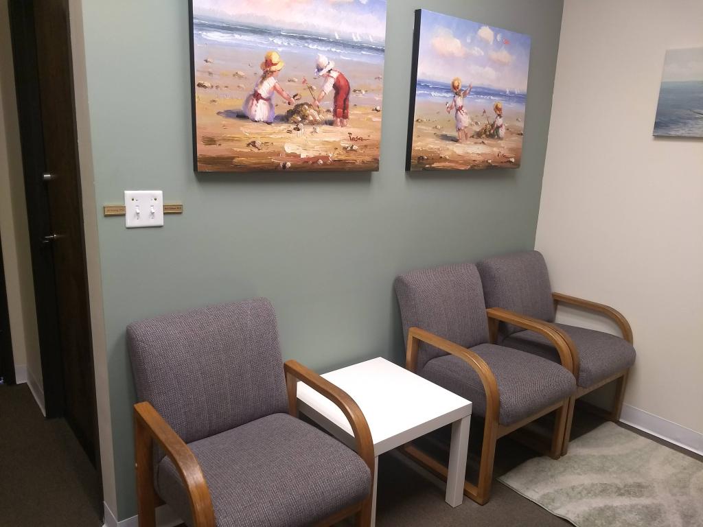Image description: Ventura office's carpeted waiting room featuring a green accent wall, 2 canvas prints of children playing on the beach, 3 chairs, and a small end table.