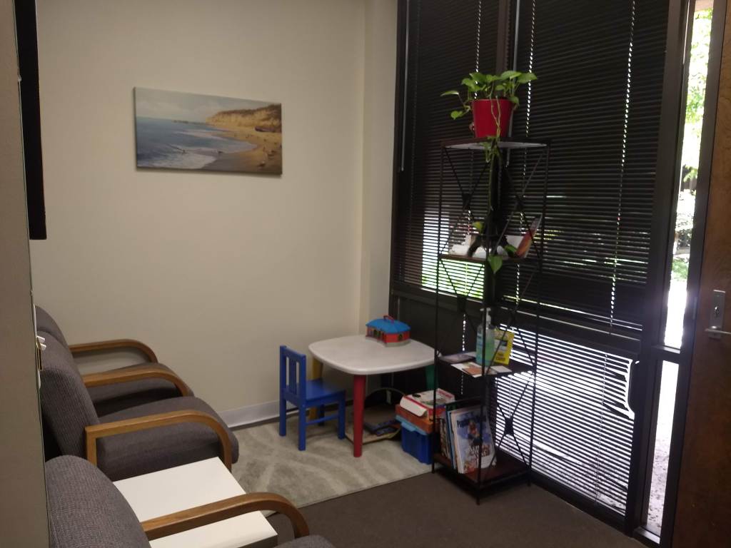 Image description: Ventura office's carpeted waiting room from a different angle featuring a children's play table, white wall, and the glass window front wall.