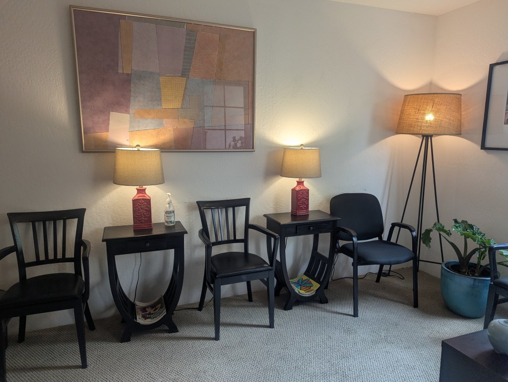 Image description: Santa Barbara office's carpeted waiting room featuring white walls, an abstract painting, 2 matching black end tables, 2 matching red table lamps, and 2 chairs.
