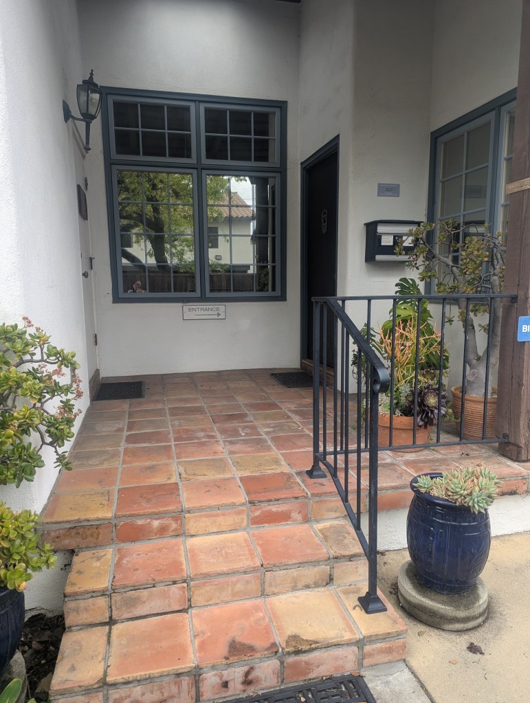 Image description: Santa Barbara office's exterior image of the front door featuring 2 steps with railings to an enclosed front patio. There's a sign that says "Entrance" with arrows pointing to the right where the wooden entrance door is. That's where you enter the office building.
